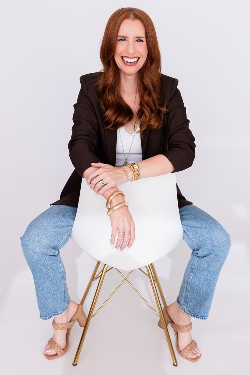 Jacqui Nelson seated on a white chair against a white studio backdrop, in a dark blazer and jeans, smiling at the camera