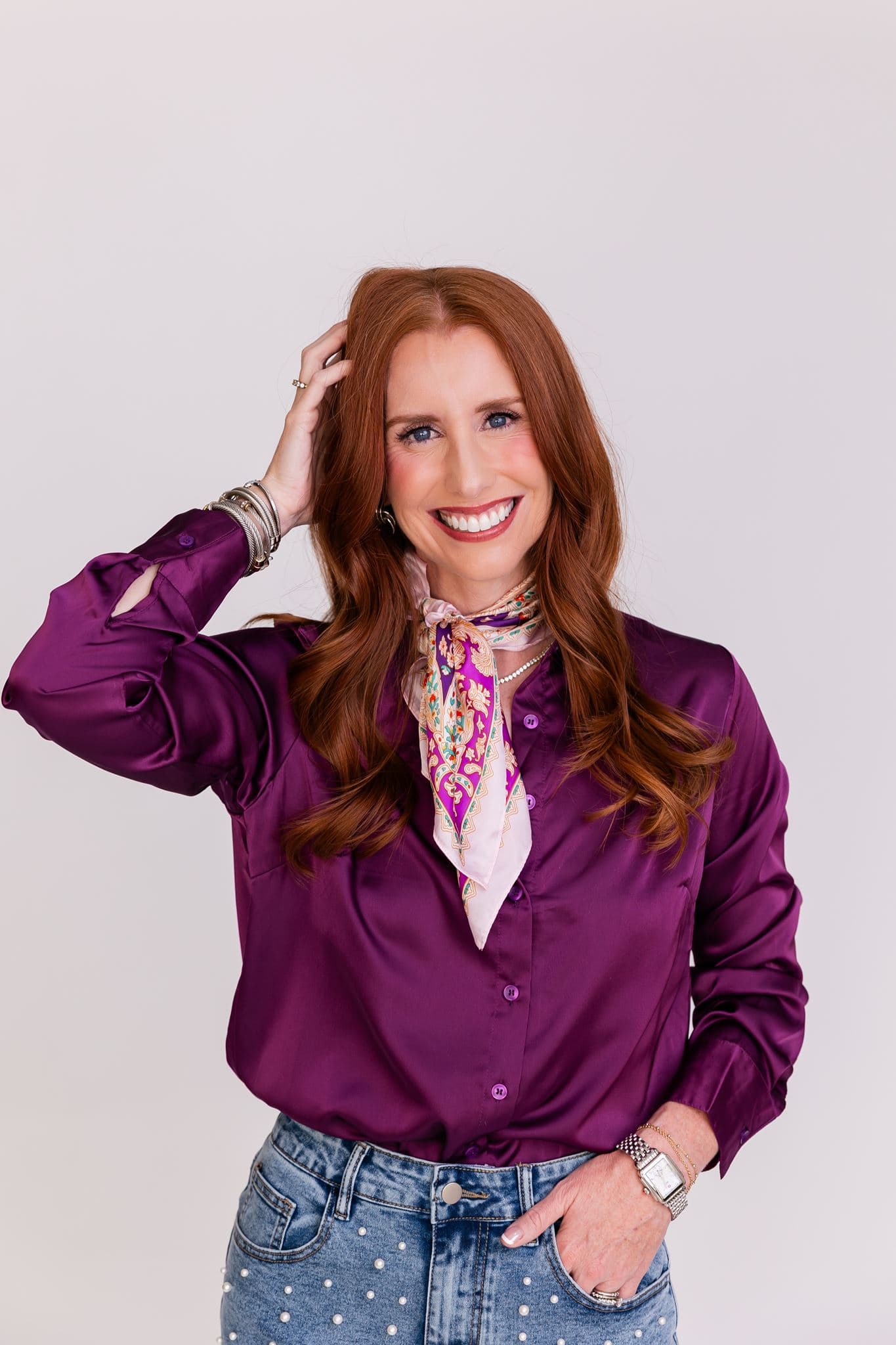Jacqui Nelson in a purple satin blouse and patterned neck scarf, one hand in her hair, smiling at the camera